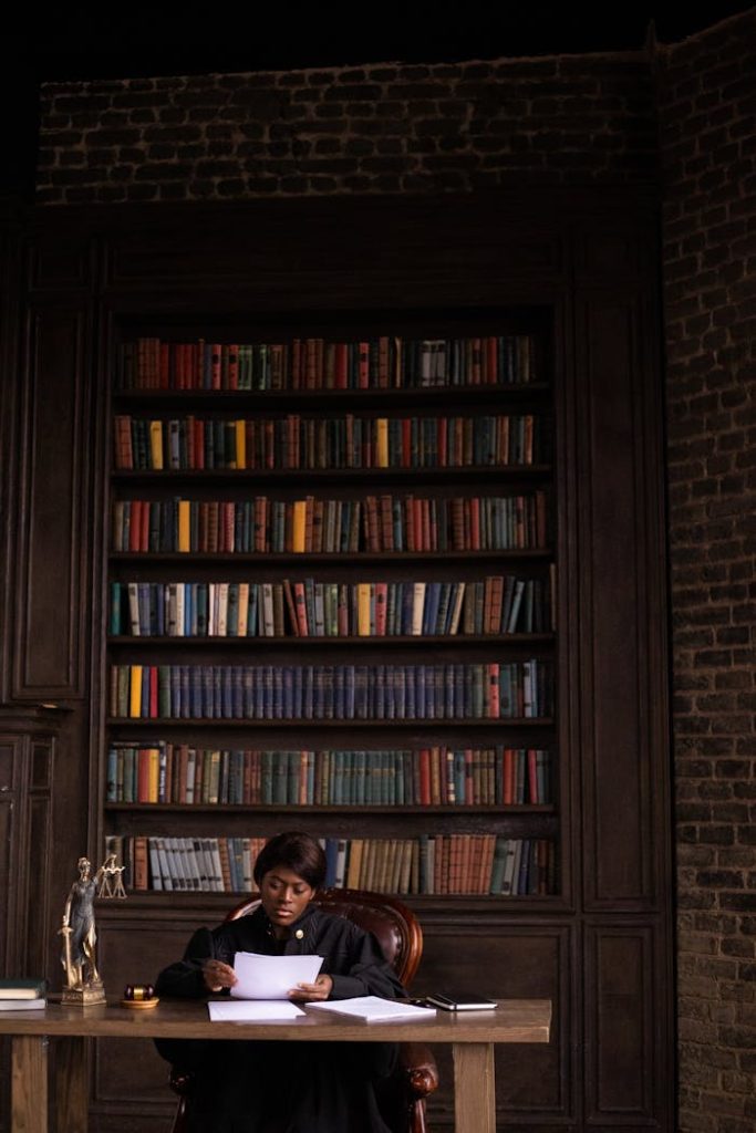 a-woman-sitting-at-the-table-6077355 Black female judge reading papers at desk in office with bookshelves.